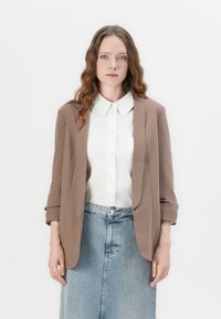 Barna blézer egy fehér galléros ing felett, világoskék farmer szoknyával viselve. Simára csiszolt textúra, laza szabás, minimalista design.