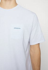 Balts kokvilnas T-krekls ar apaļu kakla izgriezumu, īsām piedurknēm un kreisajā krūtīs kabatu ar gaismas zilu "Patagonia" logotipu.