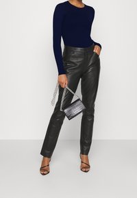 Frau trägt ein navy-blaues, geripptes Langarmoberteil, schwarze Lederhosen, schwarze Riemchenheels und hält eine kleine schwarze Handtasche mit silberner Kettenriemen.