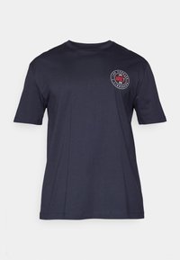 T-shirt de algodão azul-marinho com mangas curtas e gola redonda. Apresenta um logotipo circular no lado esquerdo do peito em vermelho e branco.