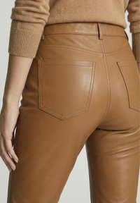 Close-up van een persoon die een hooggetailleerde, tan leren broek met achterzakken draagt, gecombineerd met een bijpassende tan gebreide trui met geribde manchetten.