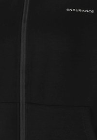 Svart zip-up jacka tillverkad av slät tyg, med en framdragare, två sidofickor och ordet "ENDURANCE" tryckt i vitt.