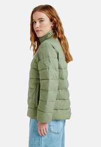 Ljusgrön puffjacka med hög krage, horisontellt quiltat mönster, med en sidficka och logotyp-etikett på vänster ärm.