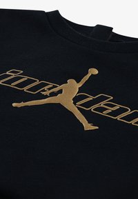 Juodas megztinis su auksiniu „Jordan“ logotipu ir silueto dizainu. Minkšta tekstūra, apvalus kaklas ir minimalistiškas stilius.