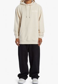 Bežová mikina s kapucí, šňůrkami, přední kapsou a logem na hrudi. V kombinaci s volnými černými kalhotami a bílými teniskami. Jednoduchý design a střih.
