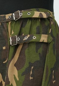 Rohelise, pruuni ja beeži toonides kamuflaažmustriga kangas, millel on vöökohal metallist pandlad ja silmused ning reljeefne pind.