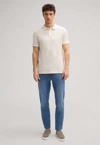 Lichtbeige polo shirt met een kraag, korte mouwen en een klein geborduurd logo. Gecombineerd met een nauwsluitende blauwe jeans en grijze instappers.
