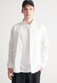 Weißes Hemd mit Knopfleiste aus Baumwolle, lange Ärmel, mit spitzem Kragen und Frontknöpfen, über einem weißen T-Shirt getragen. Schwarze Kappe auf dem Kopf.