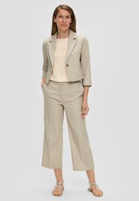 Beige linnen blazer met driekwartmouwen en een enkele knoopsluiting, gecombineerd met losse beige broeken en platte sandalen.