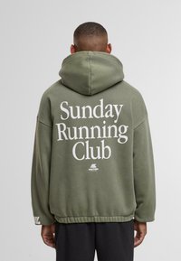 Hombre con sudadera verde oliva que tiene el texto "Club de Correr del Domingo" en la parte de atrás, de pie contra un fondo gris claro liso.