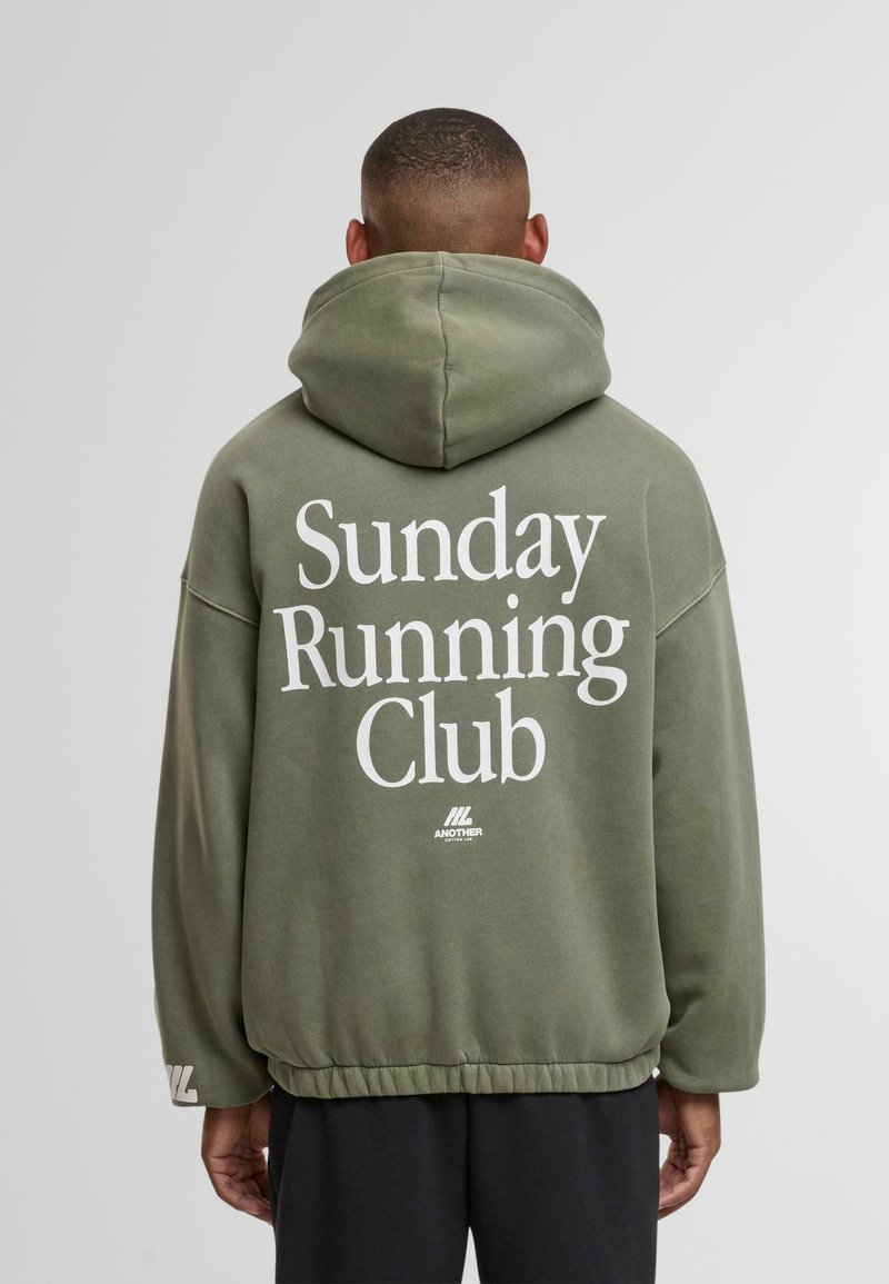 Hombre con sudadera verde oliva que tiene el texto "Club de Correr del Domingo" en la parte de atrás, de pie contra un fondo gris claro liso.