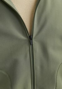 Chaqueta verde oliva con cremallera, hecha de una tela suave y texturada, con una cremallera negra y forro interior color crema, destacando su diseño funcional.