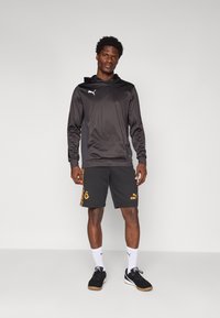 Haut de sport noir avec un logo, associé à un short gris avec des accents orange et un logo. Des baskets noires et des chaussettes blanches complètent le look.