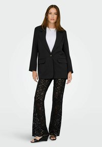 Černý blazer s klopovými límci a jedním knoflíkem, spárovaný s černými krajkovými kalhotami s širokými nohavicemi. Jednoduchý bílý top a černé klínové sandály.