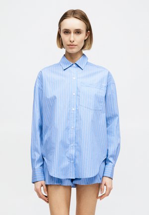 Ung kvinde med kort blondt hår iført en lyseblå, langærmet, knappet skjorte og matchende shorts mod en ensfarvet baggrund.