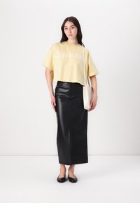 Gul croppede t-skjorte med hvit "Calvin Klein" logo, parret med en svart skinn maxi-skjørt med en frontslitt. Brukt med svarte leiligheter.