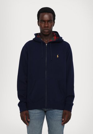 Sudadera con cremallera en color navy, con capucha forrada de cuadros, bolsillo tipo canguro y pequeño logo amarillo en el pecho. Tejido de algodón suave, corte relajado.