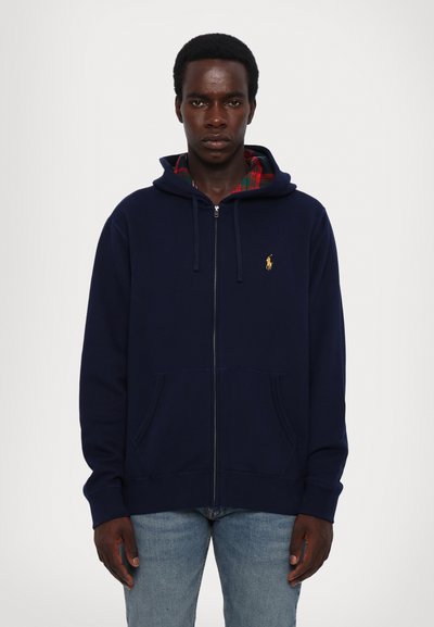 Haut à zip bleu marine avec une capuche doublée en tartan, poche kangourou et petit logo jaune sur la poitrine. Tissu en coton doux, coupe décontractée.