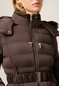 Brązowa pikowana kurtka z wysokim kołnierzem, złotymi elementami i regulowanymi troczkami. Zaprojektowana z paskiem w talii i miękką teksturą.