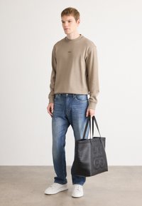 Joven con sudadera beige, jeans azules, zapatillas blancas, sosteniendo un gran bolso de cuero negro con el logo "HUGO" en relieve.