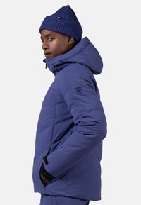 Niebieska ocieplana kurtka z kapturem, z pikowanym wzorem, zamykanymi na zamek kieszeniami oraz naszywką z logo na rękawie. Włożona z dopasowaną wełnianą czapką.