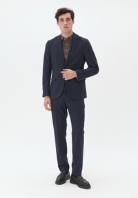 Marineblauer Anzug mit tailliertem Design, strukturiertem Stoff, einer Zweiknopfjacke und gerade geschnittenen Hosen, kombiniert mit einem braunen Hemd mit Kragen und schwarzen Schuhen.