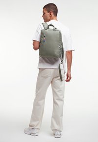 Zaino di tessuto verde con forma rettangolare, tasca frontale con cerniera e accentuazione colorata sul tirante della cerniera. È visibile la schiena di un uomo.