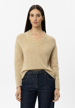 Frau mit langen dunklen Haaren, die einen beigen Pullover mit V-Ausschnitt und Spitzenbesatz sowie dunkle hoch taillierte Jeans mit schwarzem Gürtel trägt, stehend vor weißem Hintergrund.