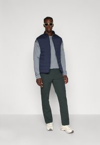 Gilet imbottito blu navy, camicia a maniche lunghe grigia, pantaloni verde scuro e scarpe da ginnastica chiare. Caratterizzato da un taglio moderno e chiusura con zip.