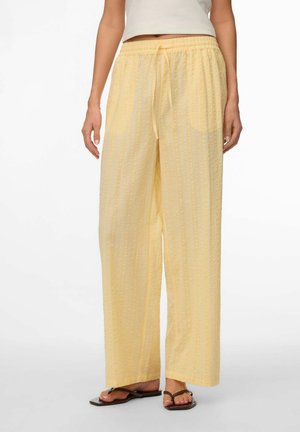 Donna che indossa pantaloni larghi di colore giallo chiaro con texture, dotati di coulisse in vita, e sandali infradito marroni, in piedi davanti a uno sfondo bianco.