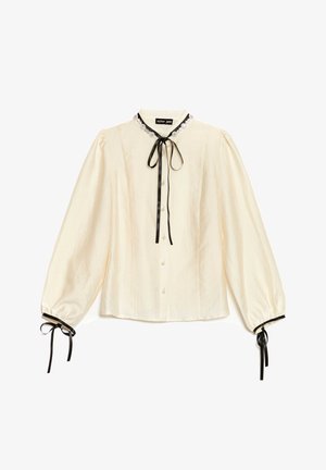 Ivoorwitte blouse met een gestructureerde afwerking, knopen aan de voorkant, zwarte lintstrik bij de kraag en zwarte accenten aan de pofmouwen met lintdetails.