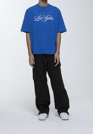 Camiseta de algodón azul con texto gráfico blanco, combinada con pantalones cargo negros holgados y zapatillas blancas. Ajuste sencillo y relajado.
