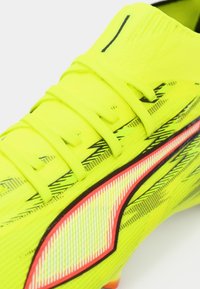 Neonowo żółty but piłkarski z czarnymi akcentami i teksturowaną cholewką, z wyraźnym czerwonym paskiem i tradycyjnym sznurowaniem.