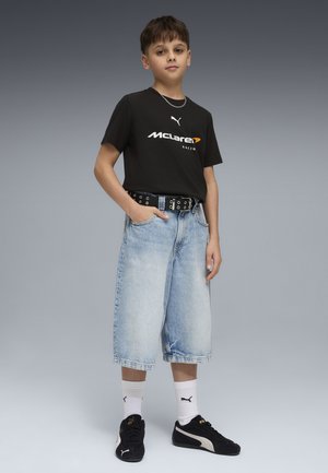 Chłopiec ubrany w czarną koszulkę McLaren Racing, jasnoniebieskie dżinsowe spodenki do kolan, białe skarpetki Puma i czarne buty sportowe Puma, stojący z jedną ręką w kieszeni.