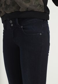 Mörka denimjeans med fem fickor, två framknappar och slim fit. Materialet har en lätt struktur och en subtil glans.