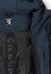 Donkerblauwe en zwarte jas met een binnenkant waarop een "TRAVELIN'" logo patch en geborduurde tekst "waar we lopen" is te zien, geopend om de voering te tonen.