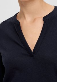 Marinblå topp med V-ringning, tillverkad av mjukt tyg med en slät textur. Minimalistisk stil, utan synliga mönster eller detaljer.