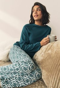 Türkiser Langarm-Oberteil kombiniert mit gemusterten, fließenden Hosen im grünen und weißen Blumenmuster. Vor einem gemütlichen, strukturierten, beigen Hintergrund.