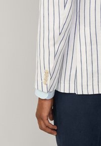 Blazer en lin blanc avec des rayures verticales bleues, présentant un design à un seul bouton, quatre fermetures à bouton et un poignet de chemise bleu clair visible en dessous.