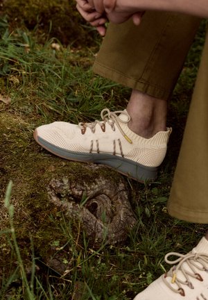 Personne portant des baskets en maille beige avec des accents marron et jaunes, marchant sur un sol mousseux avec de l'herbe et une racine d'arbre visibles.