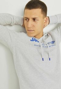 Sudadera ligera de color gris claro hecha de un tejido suave, con un logo azul y cordones con puntas azules. El diseño incluye un ajuste relajado y un bolsillo tipo canguro.