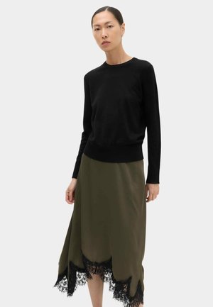 Vrouw die een zwarte top met lange mouwen en een asymmetrische olijfgroene rok met zwarte kanten zoom draagt, staand tegen een lichte achtergrond.