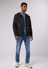 Svart huva jacka med två framfickor, bärs över en blå skjorta, kombinerad med ljusblå jeans och neutralt färgade sneakers.