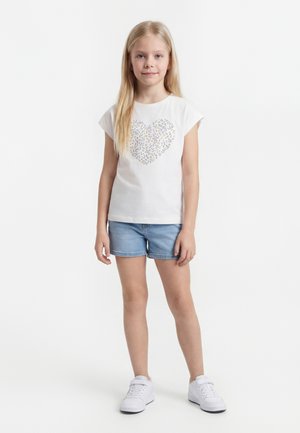 Noor tüdruk pikkade blondide juustega, kes kannab valget särki lillemustrilise südamega, teksapükse ja valgeid tenniseid, seisab valgel taustal.