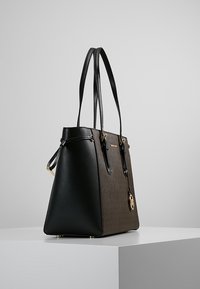 Zwarte en bruine neplederen schoudertas met lange banden, textuurafwerking, goudkleurige hardware en een decoratieve logo-hanger.