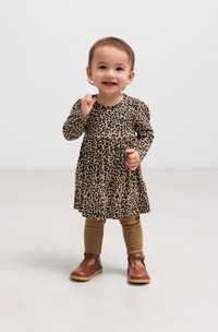 Småbarn smiler mens han står på et lyst gulv, iført en kjole med leopardmønster, brune leggings og brune sko.