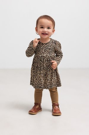 Småbarn smiler mens han står på et lyst gulv, iført en kjole med leopardmønster, brune leggings og brune sko.