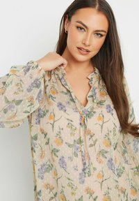 Blusa floreale in tessuto leggero e trasparente con una base beige. Presenta fiori pastello, scollo a V con bordino arricciato e maniche lunghe a sbuffo.