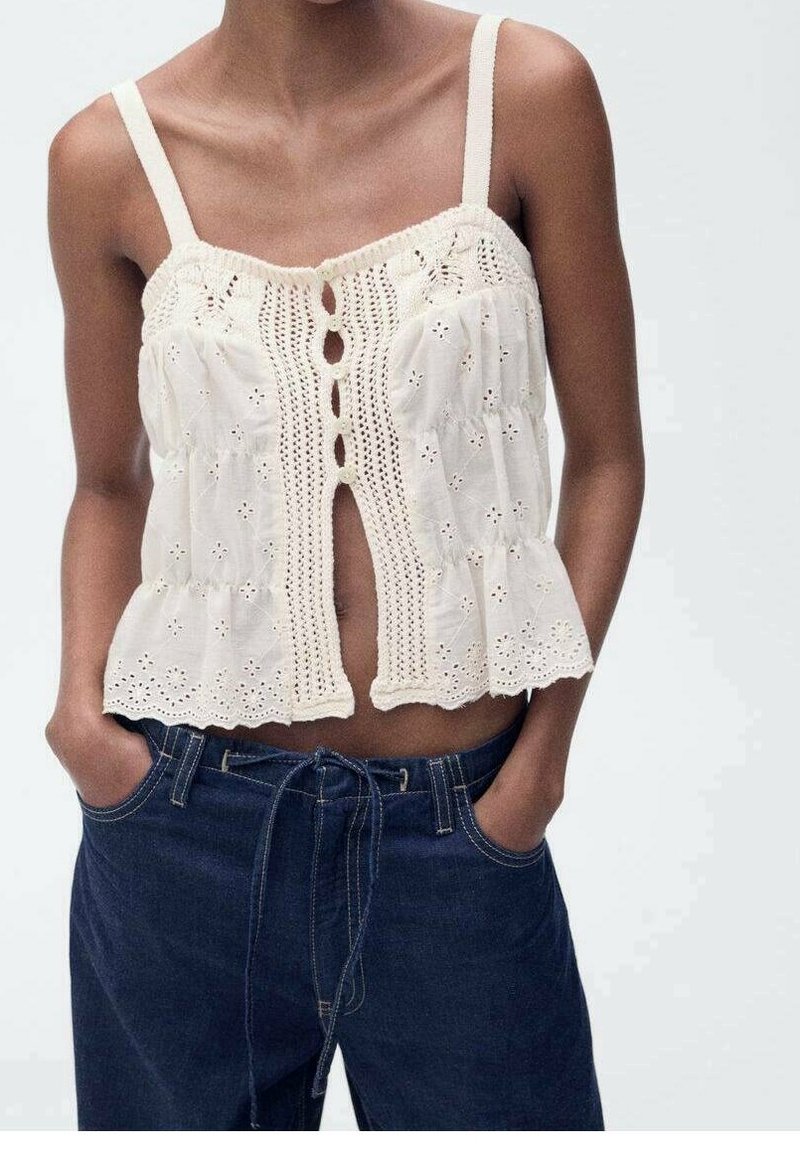 Femme portant un haut blanc crocheté sans manches avec des découpes florales et un jean bleu foncé taille haute, les mains dans les poches.