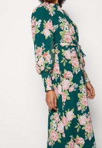 Vestido floral con una base verde y rosas rosas, mangas largas con fruncido, cuello alto y un cinturón atado en la cintura para una silueta definida.